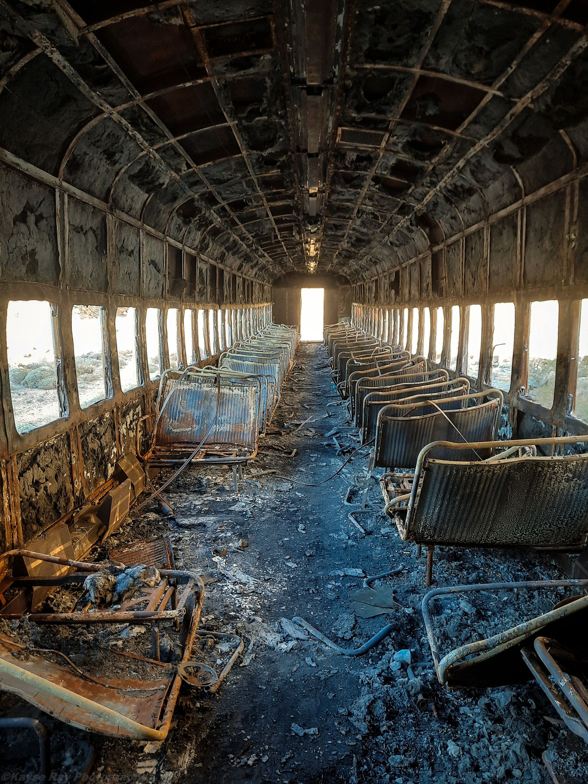 Burned-out interior of an abandoned passenger rail car with charred seats leading to a bright doorway
