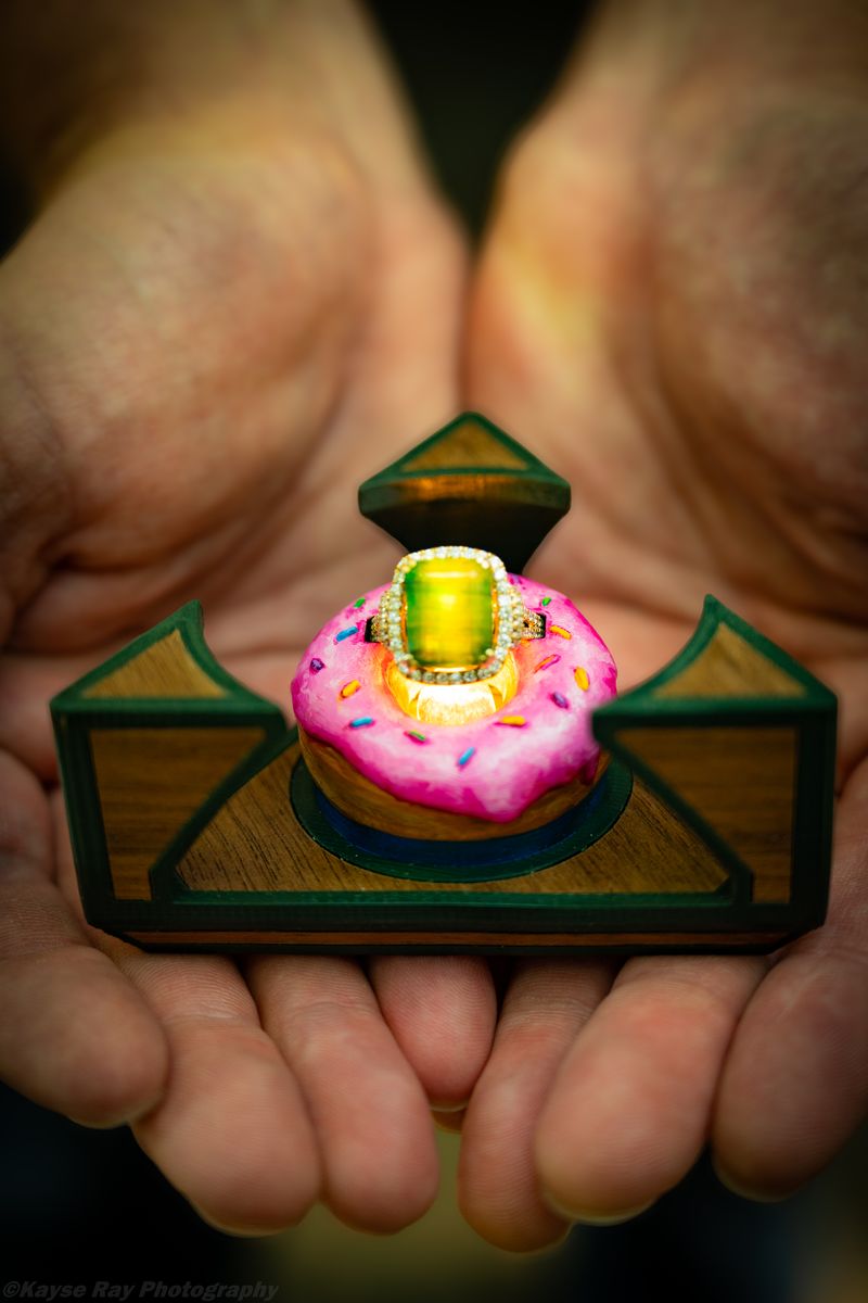 Cupped hands hold a wooden ring box shaped like a pink sprinkled donut cradling a glowing emerald ring.