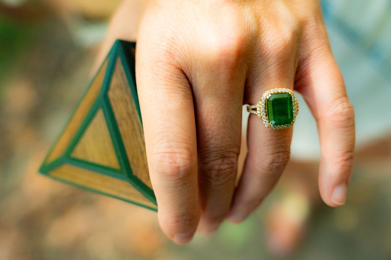 Close-up of an emerald and diamond halo engagement ring on a hand beside a triangular wooden ring box.