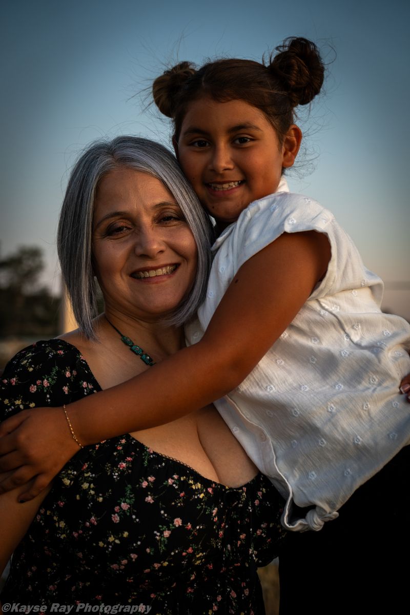 A silver-haired grandmother smiles while embracing her granddaughter against a soft dusk sky.