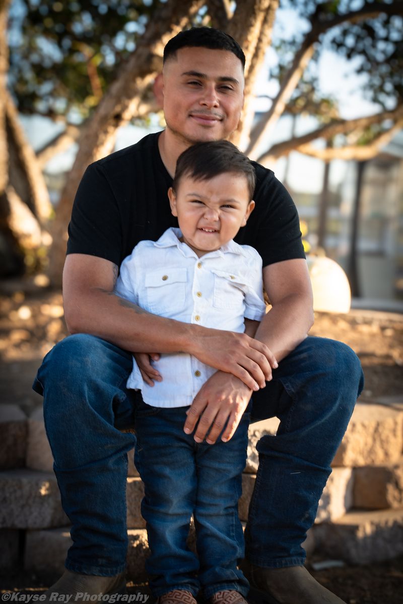 A father sits on stone steps in golden-hour light holding his young son, who scrunches his face in a grin.