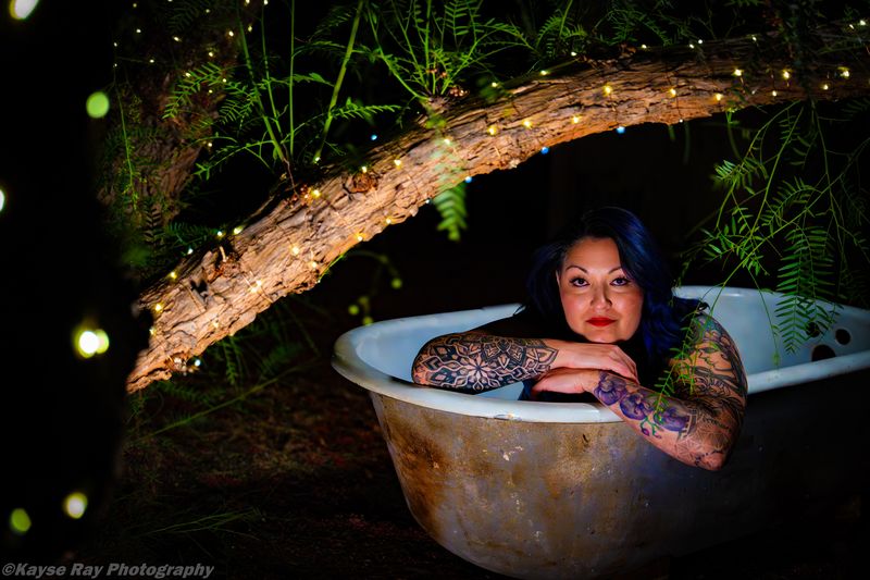 Blue-haired woman with tattooed arms resting her chin on crossed arms at the edge of a rustic clawfoot tub beneath a fairy-lit branch.