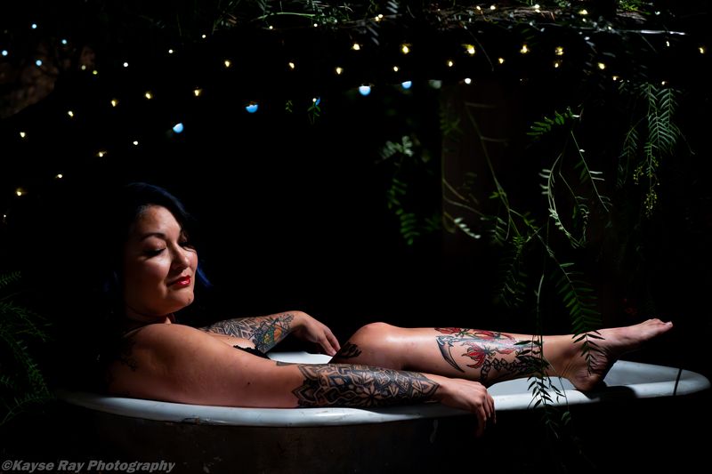 Tattooed woman reclining serenely in a clawfoot tub with her feet on the rim under a canopy of twinkle lights.