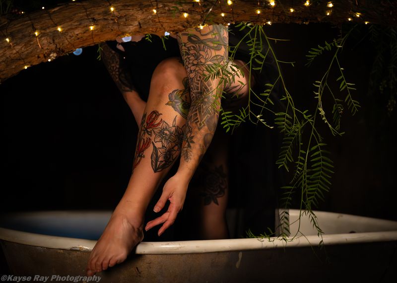 Detail of floral leg tattoos and a graceful hand draped over a tub rim beneath a light-wrapped branch.