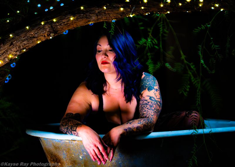 Blue-haired woman leaning on the edge of a rustic tub, gazing down pensively under strings of fairy lights.