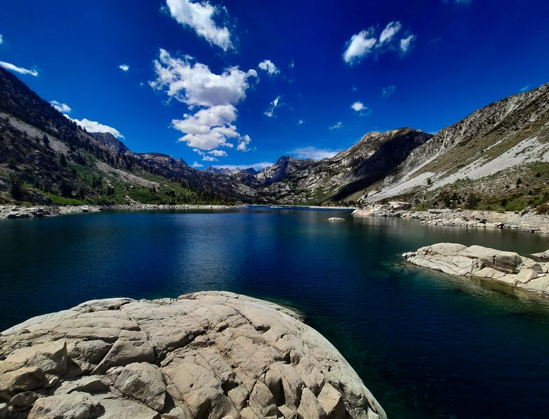 Deep blue alpine lake ringed by granite peaks under a bright sky, with a pale rock slab in the foreground