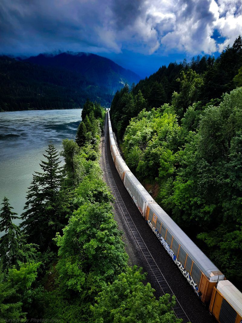Freight train curving along a river gorge beneath moody storm clouds and mist-covered mountains